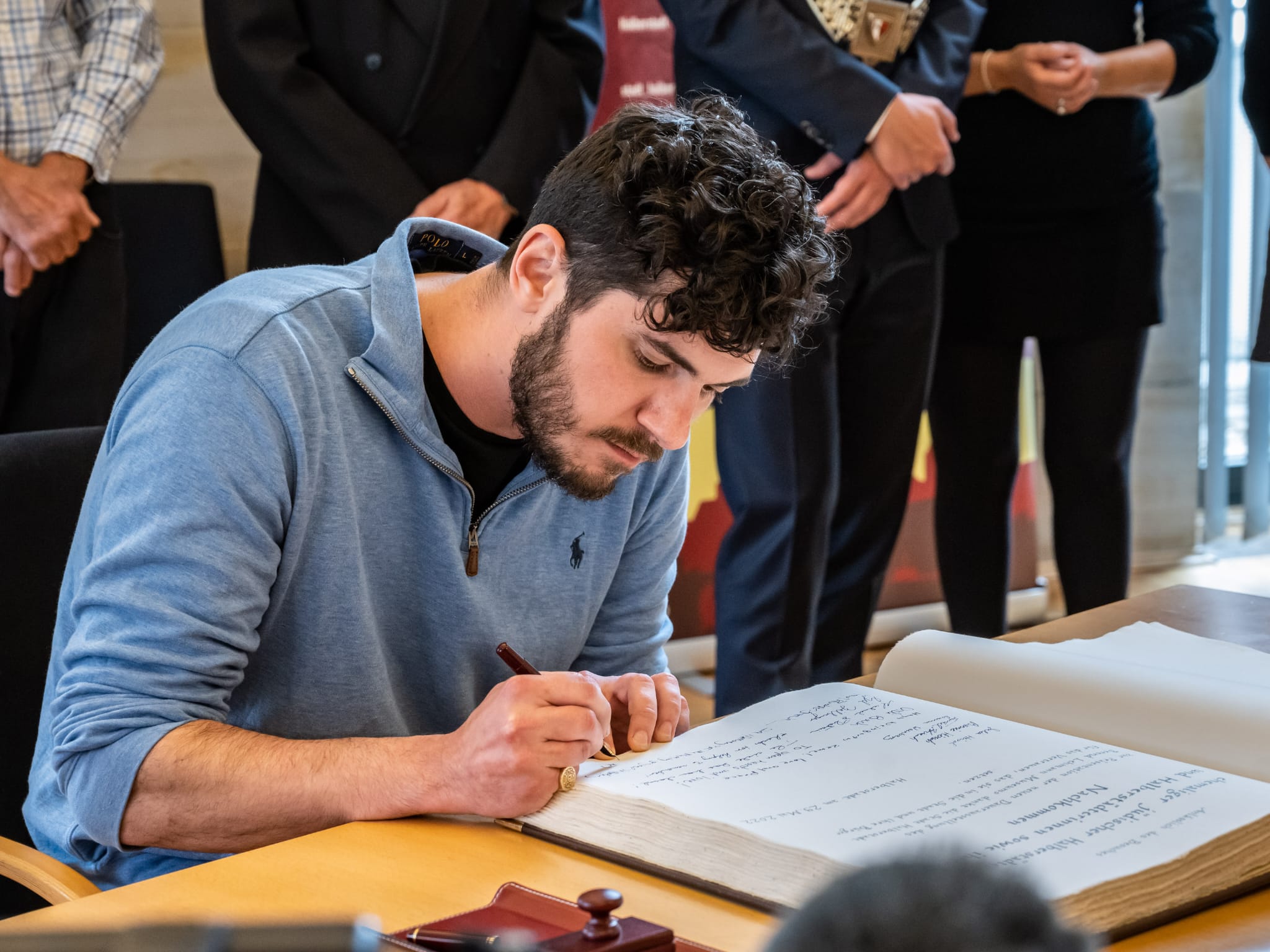 Rabbi Daniel Dolph Reichenbach seated at a town-hall table, writing in Halberstadt’s open Golden Book while other attendees stand behind him